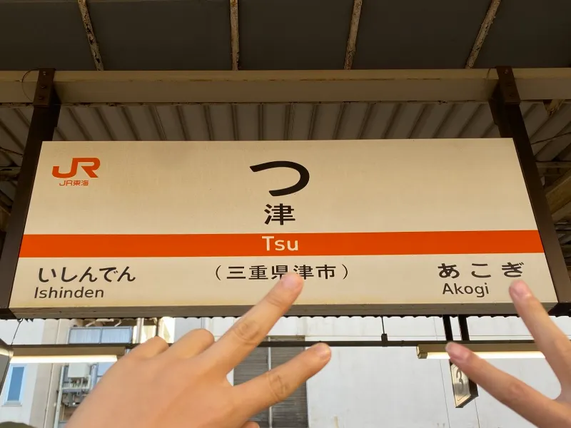 津駅の駅名看板の前でピースする成人男性の画像