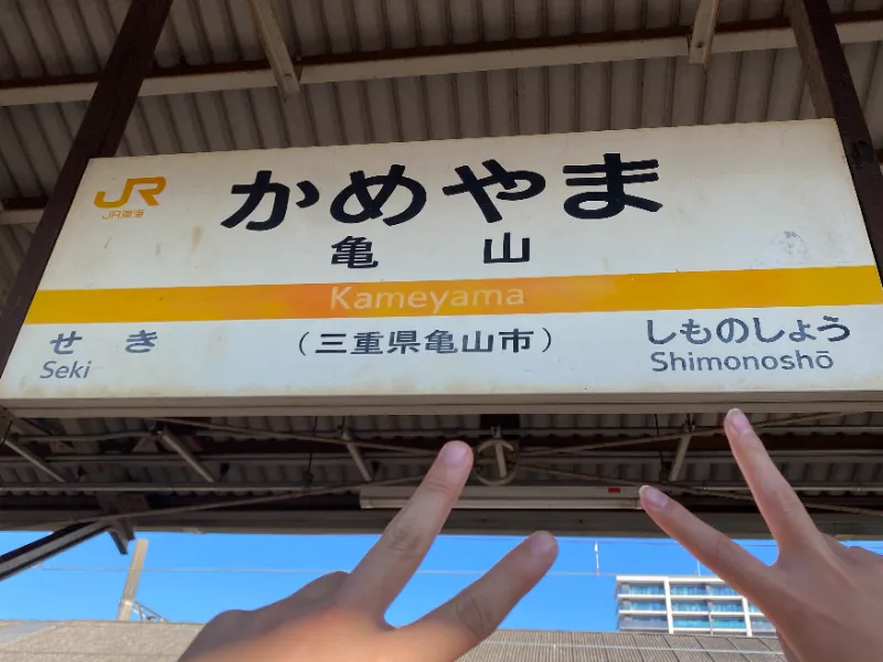 亀山駅の駅名看板の前でピースする成人男性の画像。