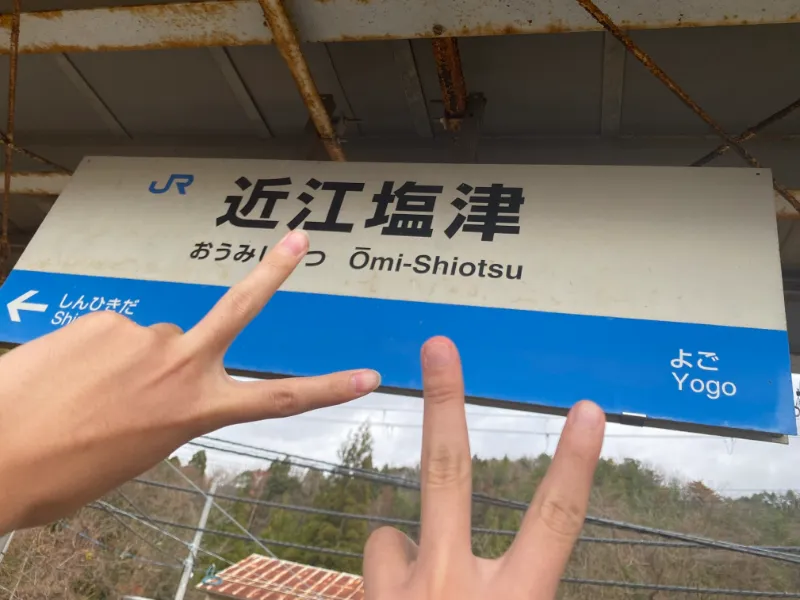 近江塩津駅の駅名看板の前でピースする成人男性の画像。
