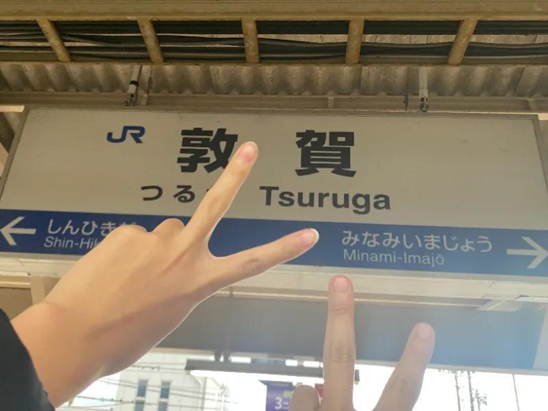 敦賀駅の駅名看板の前でピースする成人男性の画像。