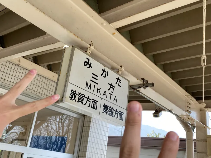 三方駅の駅名看板の前でピースする成人男性の画像。