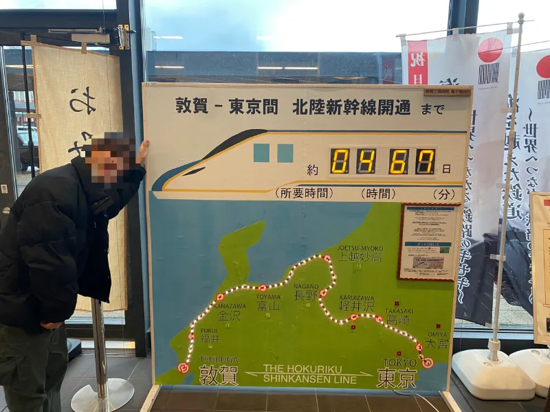 北陸新幹線沿線までのカウントダウンが表示されている看板の前で笑う成人男性の画像。